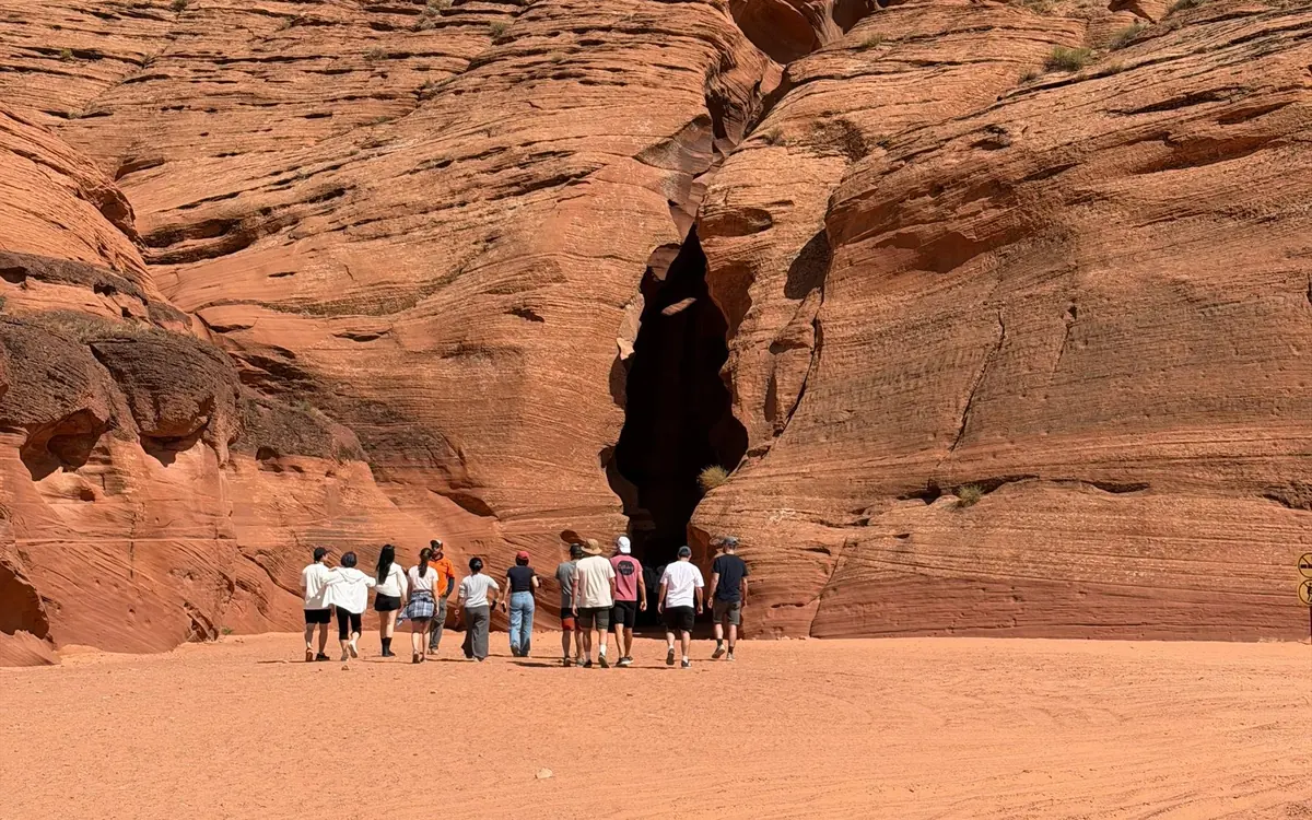 Eine Gruppe läuft über offenen Sand auf einen dunklen Spalt in einer Sandsteinwand zu
